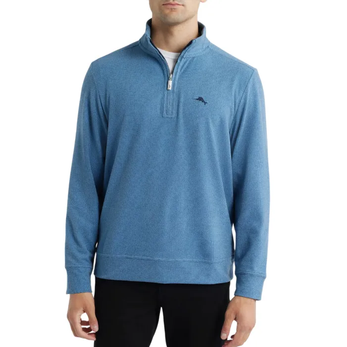 A man is wearing a blue quarter-zip pullover sweater with a small embroidered logo on the chest, paired with black pants.