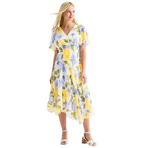 A woman wears a floral wrap dress featuring yellow and blue hues, knee-length with short sleeves, paired with white sandals.