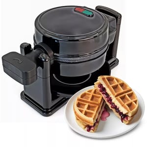 A black, rotating Belgian waffle maker with two indicator lights on top, alongside a plate with a waffle sandwich filled with berries and cream.