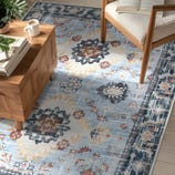 A wooden coffee table with a cup of coffee and a stack of books sits on a blue, patterned rug. An upholstered chair with wooden legs and an open book is nearby.