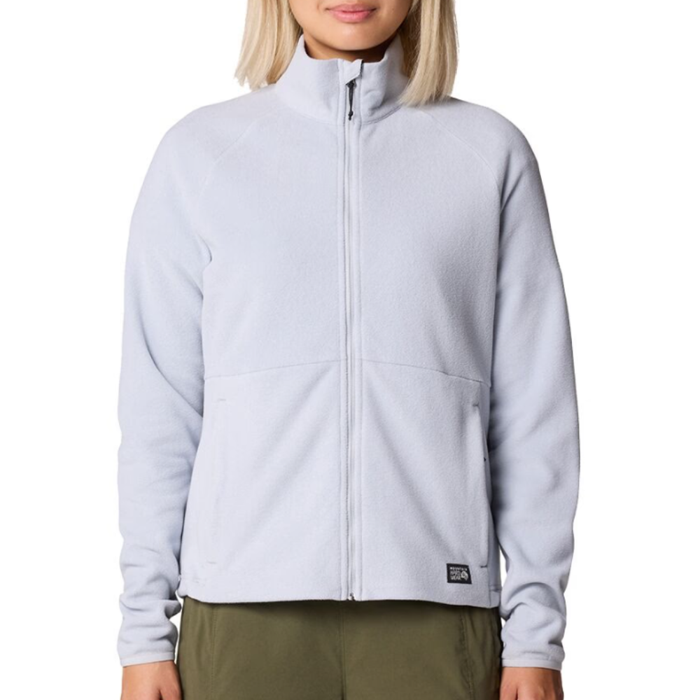 A woman is wearing a light gray zip-up fleece jacket with a high collar and a logo tag at the hem. The jacket features long sleeves and is paired with olive green pants.