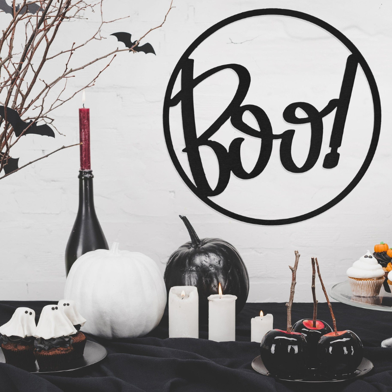 A Halloween-themed setup with cupcakes, black and white pumpkins, candied apples, candles, and a decorative \“Boo!\“ sign, surrounded by branches and bat cutouts.