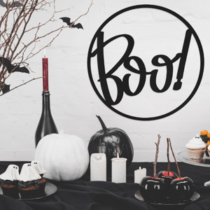 A Halloween-themed setup with cupcakes, black and white pumpkins, candied apples, candles, and a decorative \“Boo!\“ sign, surrounded by branches and bat cutouts.
