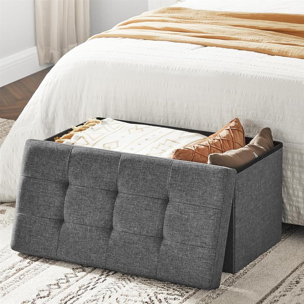 Gray upholstered storage bench at the foot of a white bed, partially open revealing pillows and blankets.