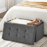 Gray upholstered storage bench at the foot of a white bed, partially open revealing pillows and blankets.