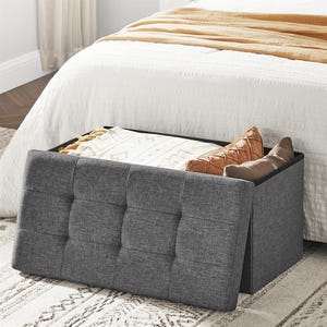 Gray upholstered storage bench at the foot of a white bed, partially open revealing pillows and blankets.