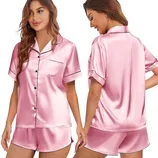 Women's satin pajama set in pink featuring a short-sleeve button-up top with a chest pocket and matching shorts, accented with black piping.