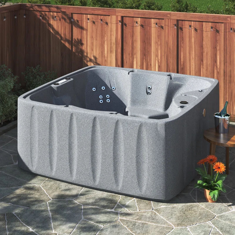 6-Person Waterfall Hot Tub with a grey, textured exterior features multiple jets for hydrotherapy. It's placed outdoors on a stone patio, accompanied by a small table with a champagne bucket and flowers nearby.