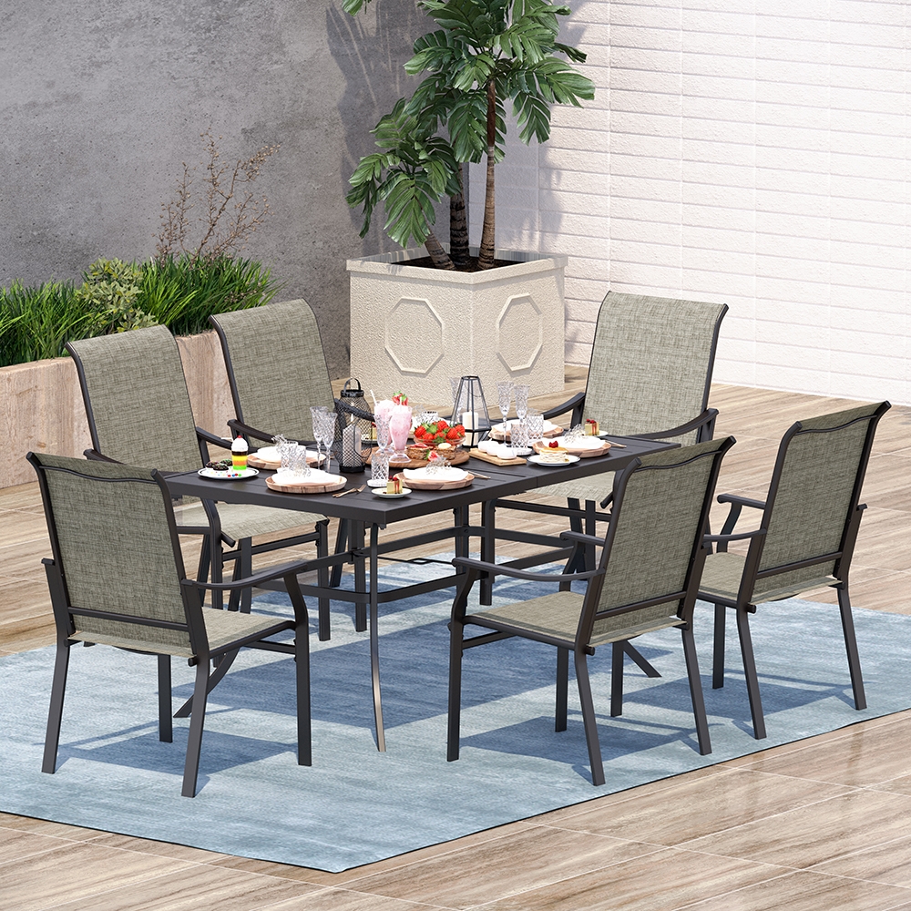 The 7pc Patio Dining Set features a rectangular black table with six green cushioned chairs, ideal for outdoor dining. It rests on a light blue rug, surrounded by plants, and is set with dishes, glasses, and decorative items.