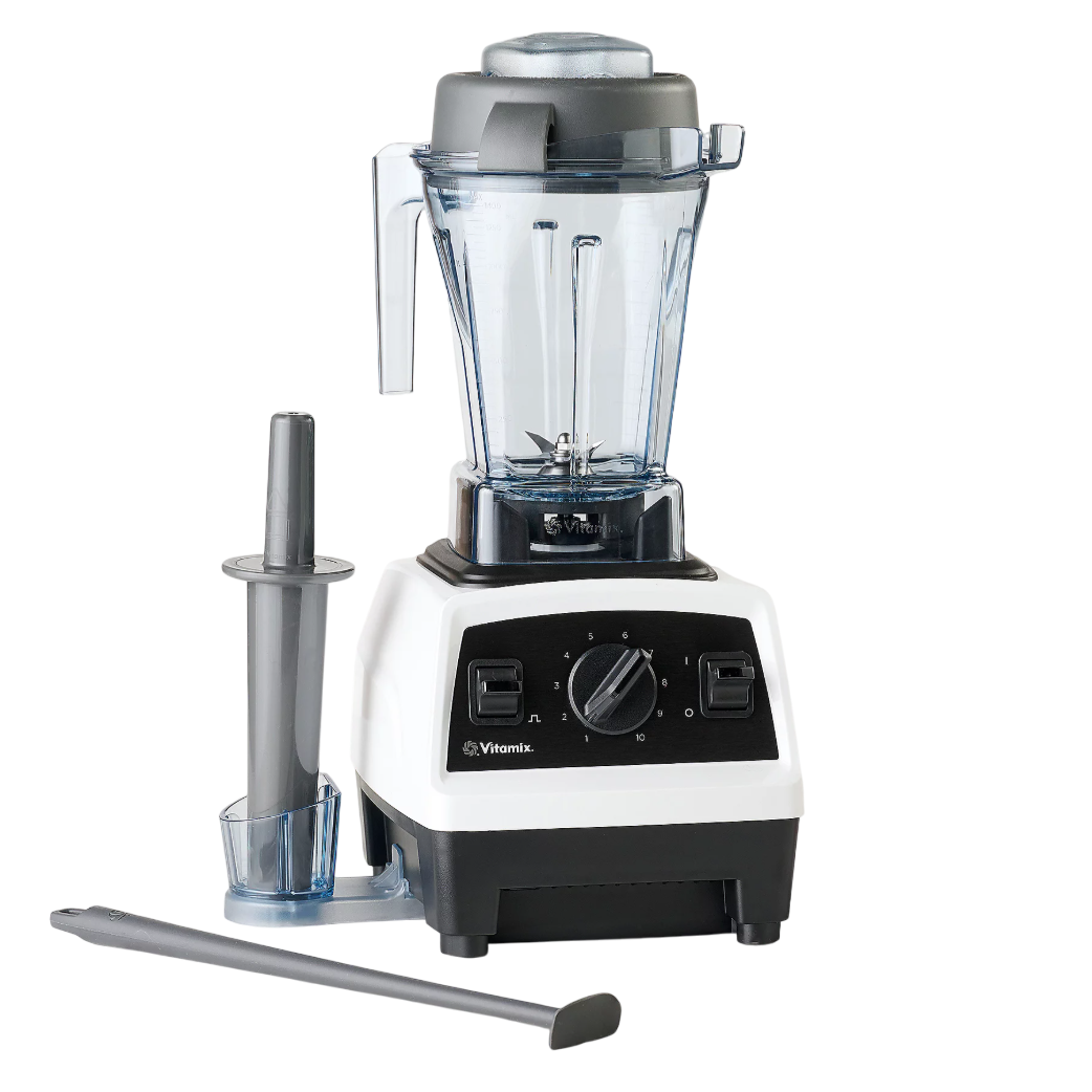 A Vitamix blender with a clear pitcher, black dial, and white base, accompanied by a tamper and a scraper tool, is shown on a light-colored kitchen counter.