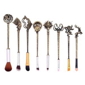 Eight makeup brushes with ornate handles, each featuring symbols from \“Game of Thrones\“ houses. The set includes brushes for various cosmetic applications with intricate designs in a bronze finish, representing themes from the series.