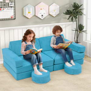 Two children are sitting on a convertible blue play couch with footrests, reading books. The couch can be configured in different ways and is surrounded by playful wall decor and a potted plant.
