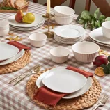 A dining table setup with a checkered tablecloth features round wicker placemats, white dinnerware, red fabric napkins, gold flatware, and a centerpiece with pears, bread, and a candle.