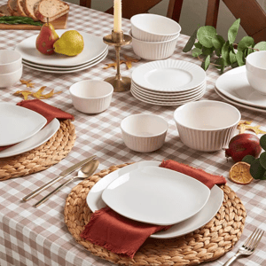 A dining table setup with a checkered tablecloth features round wicker placemats, white dinnerware, red fabric napkins, gold flatware, and a centerpiece with pears, bread, and a candle.