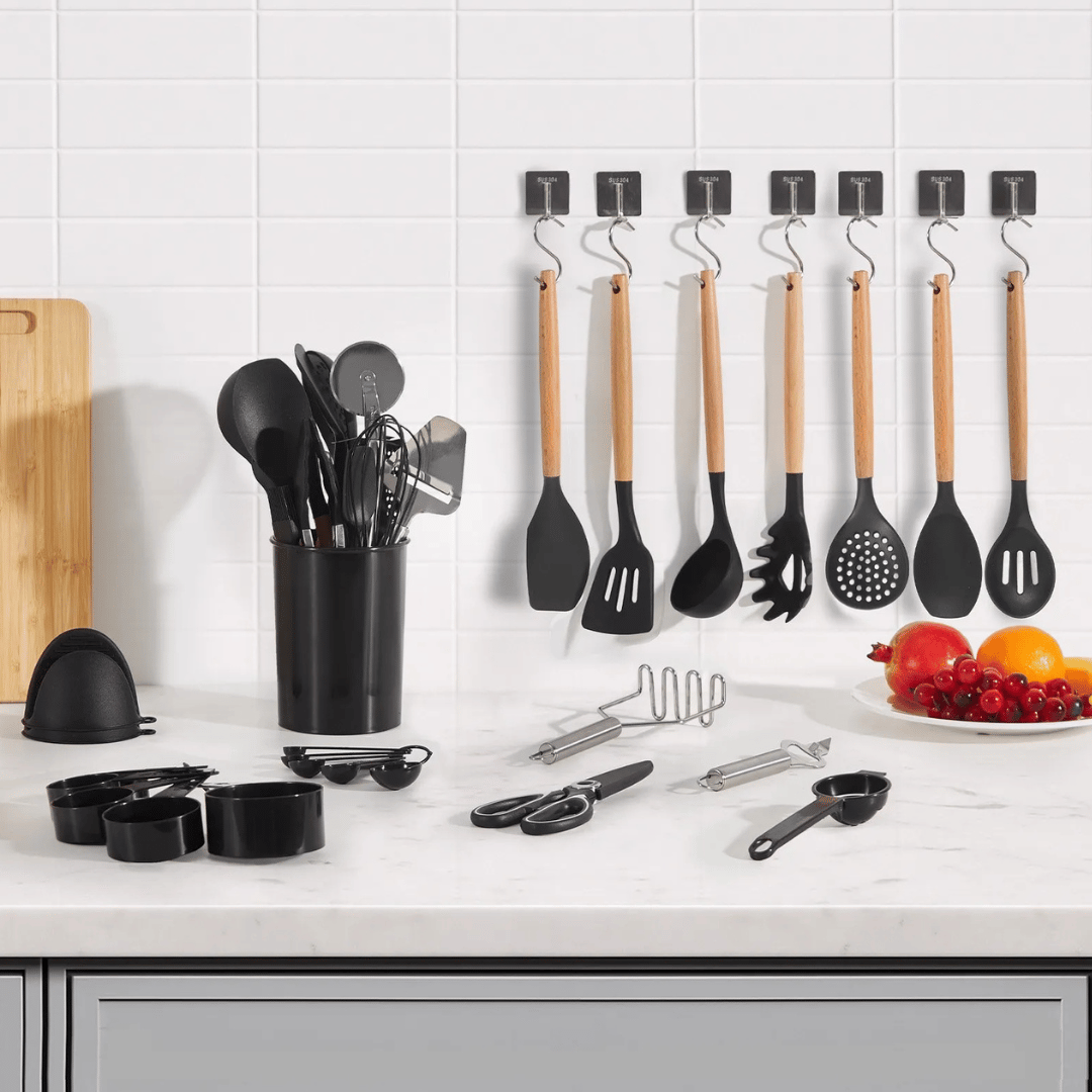 A set of kitchen utensils including spatulas, spoons, and a whisk are displayed on a countertop, with some hanging on wall hooks. Additional items include measuring cups, scissors, a peeler, a masher, and a plate of fruit.