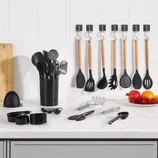 A set of kitchen utensils including spatulas, spoons, and a whisk are displayed on a countertop, with some hanging on wall hooks. Additional items include measuring cups, scissors, a peeler, a masher, and a plate of fruit.