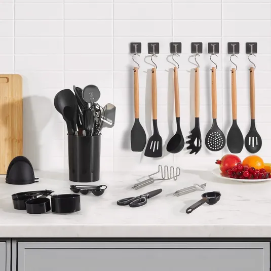 A set of kitchen utensils including spatulas, spoons, and a whisk are displayed on a countertop, with some hanging on wall hooks. Additional items include measuring cups, scissors, a peeler, a masher, and a plate of fruit.