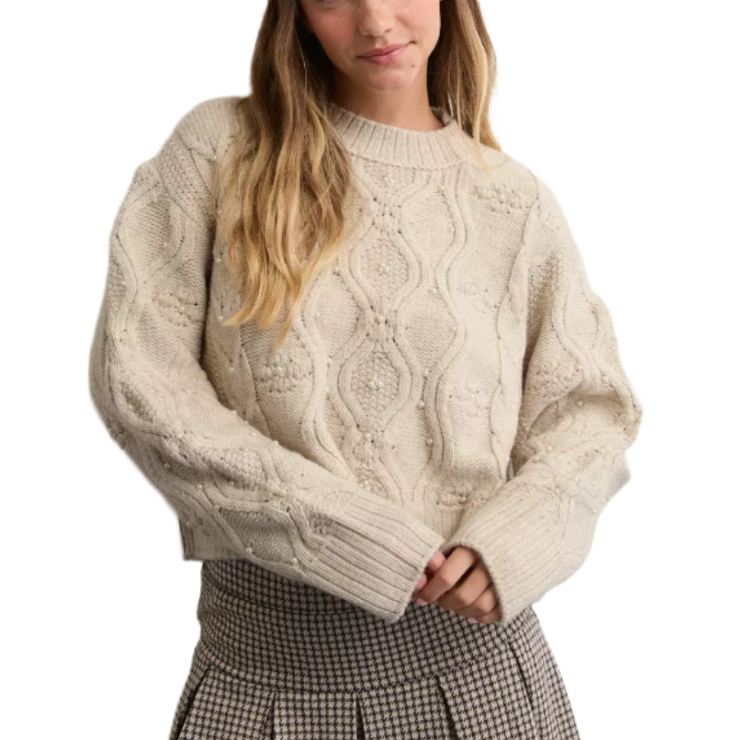 A woman wears a cream-colored, textured cable knit sweater with long sleeves and a plaid pleated skirt.