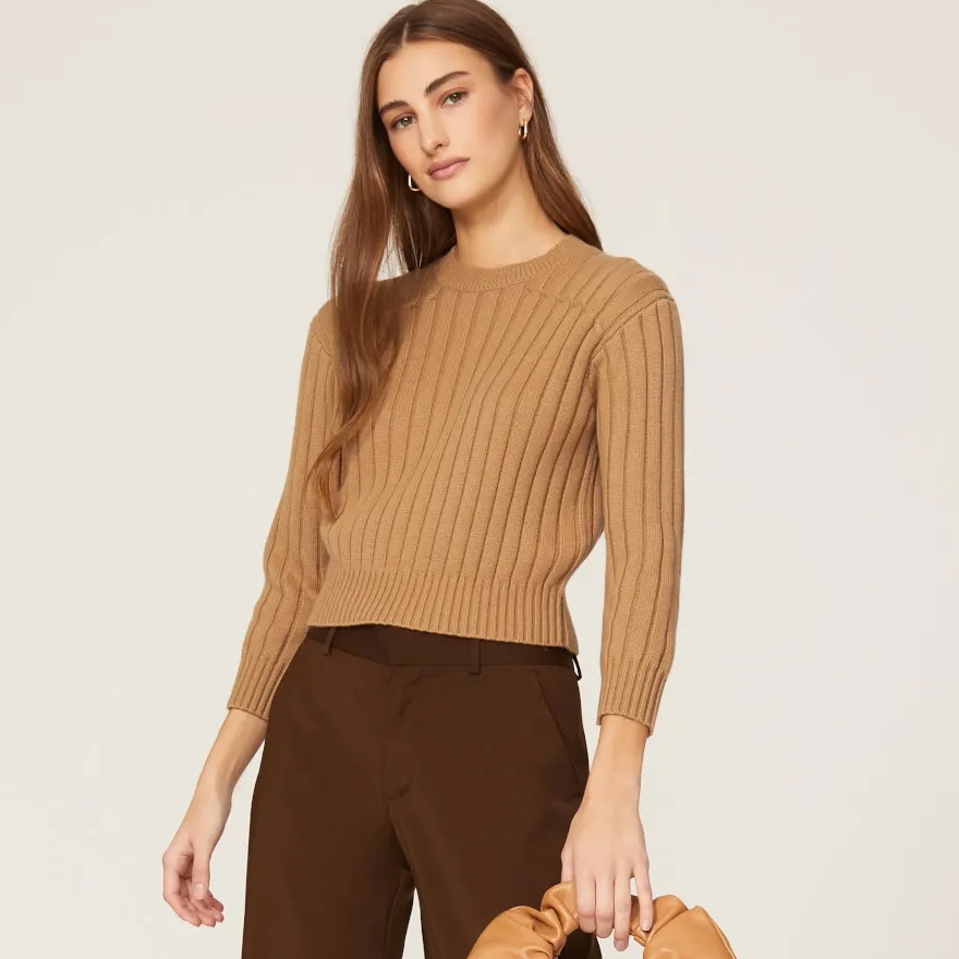Woman wearing a tan ribbed sweater and dark brown trousers, accessorizing with gold earrings.