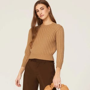 Woman wearing a tan ribbed sweater and dark brown trousers, accessorizing with gold earrings.