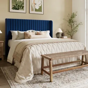 A bedroom features a blue tufted headboard, a cream quilted bedspread with beige accents, wooden nightstands with lamps, a woven bench, floral paintings, and a leafy plant by a large window.
