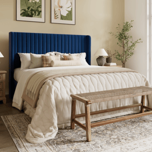 A bedroom features a blue tufted headboard, a cream quilted bedspread with beige accents, wooden nightstands with lamps, a woven bench, floral paintings, and a leafy plant by a large window.