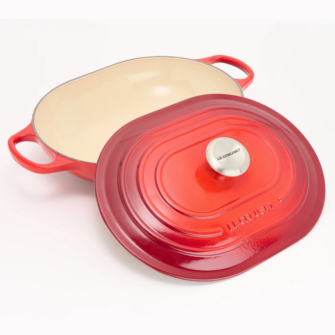 A red Le Creuset cast iron casserole dish with an oval shape, featuring an off-white interior and a stainless steel knob on the lid.