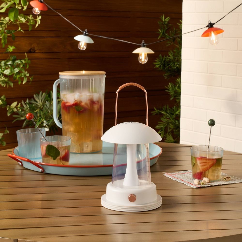 A table setting features a lantern with a brown handle, a tray with a pitcher of iced beverage containing strawberries, and two glasses with drink stirrers. Hanging string lights illuminate the outdoor scene.