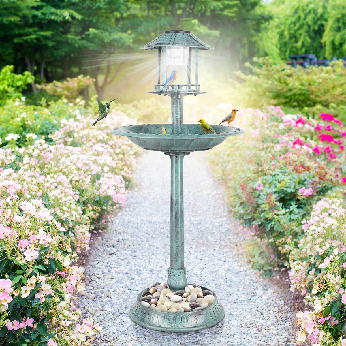 A decorative bird bath with a solar-powered lantern, adorned with various small birds, is set on a garden path lined with blooming flowers.