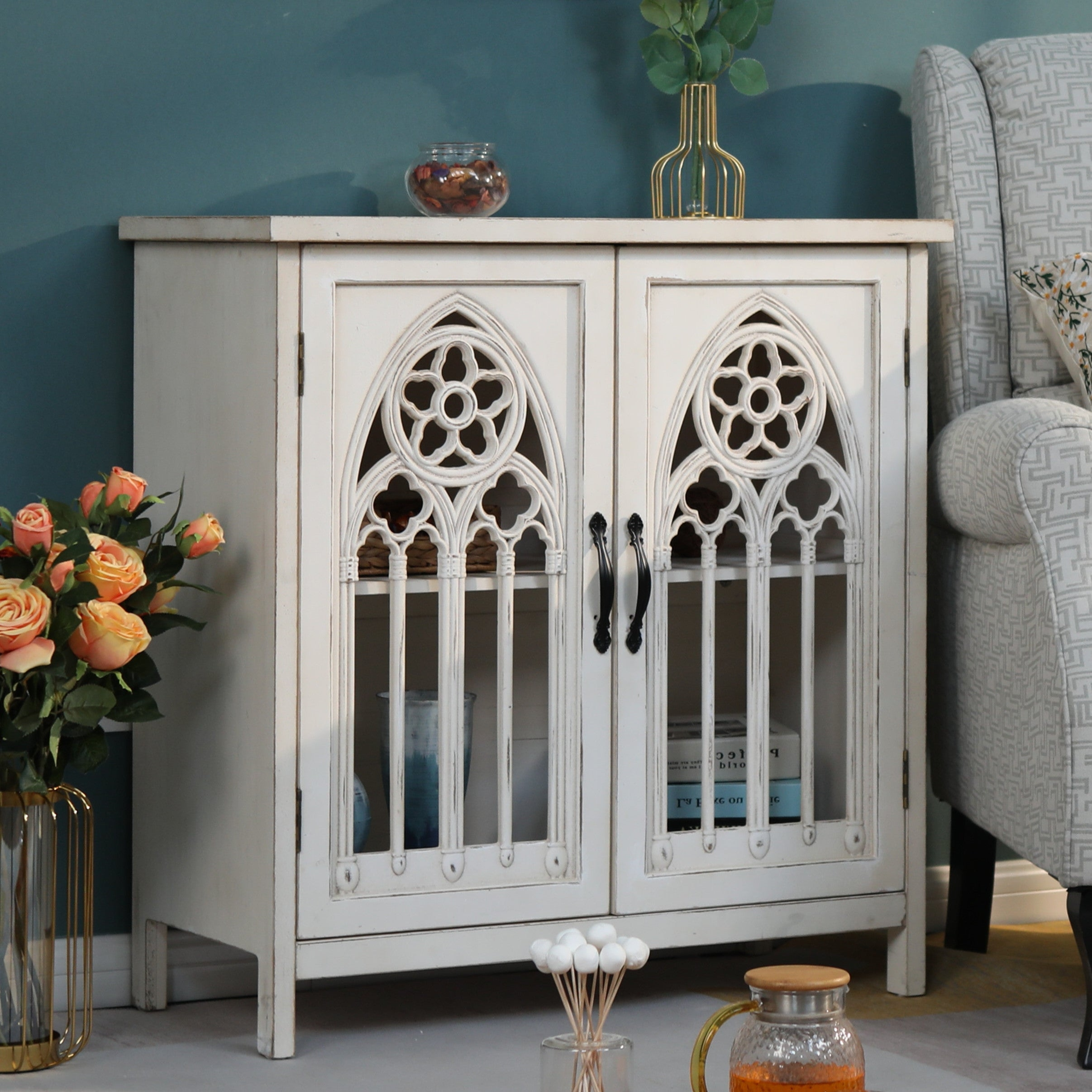 A white cabinet with Gothic-style lattice doors, featuring book and vase storage, is placed next to a gray armchair on a light floor. Nearby are a vase with roses and a glass pitcher with orange liquid.