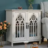 A white cabinet with Gothic-style lattice doors, featuring book and vase storage, is placed next to a gray armchair on a light floor. Nearby are a vase with roses and a glass pitcher with orange liquid.