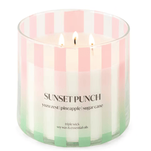 A triple wick candle labeled \“Sunset Punch\“ with notes of yuzu zest, pineapple, and sugar cane, in a glass container featuring pink, white, and green vertical stripes. Made from soy wax and essential oils.