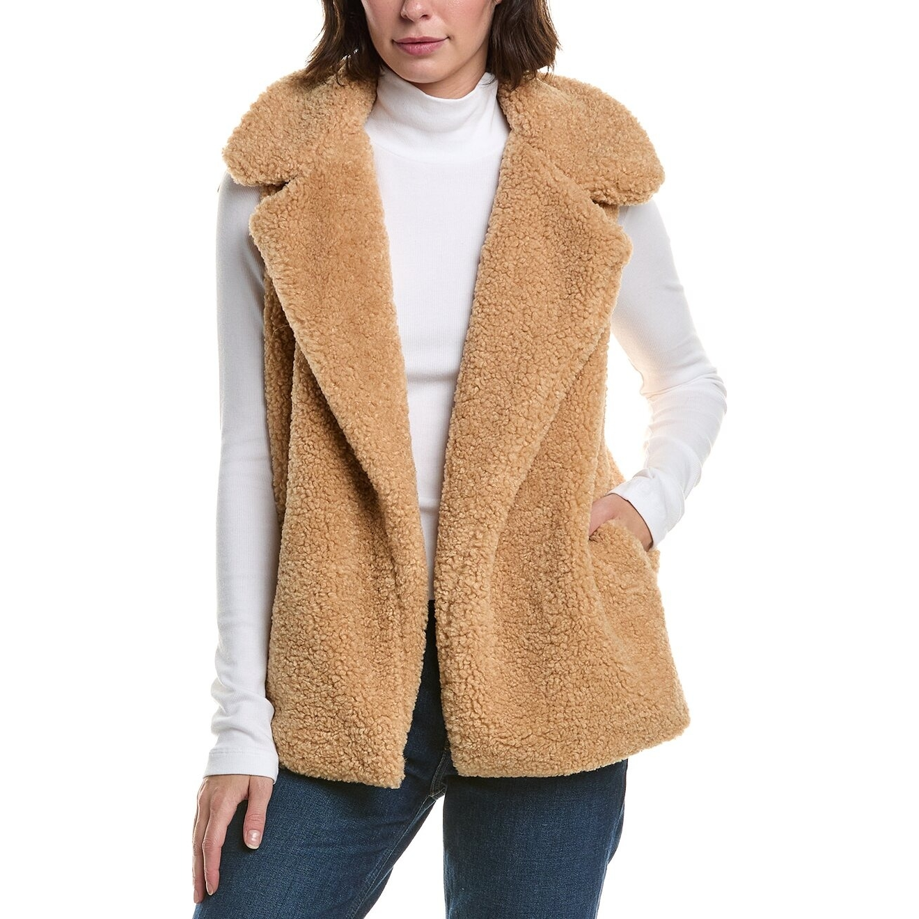 A woman is wearing a camel-colored sherpa vest over a white turtleneck with blue jeans. The vest features side pockets and a wide collar.