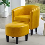 Yellow barrel chair and matching ottoman with black legs, placed on a light rug in a bright room with a window view of a garden, next to a small white side table with a lamp and a cup.