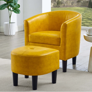 Yellow barrel chair and matching ottoman with black legs, placed on a light rug in a bright room with a window view of a garden, next to a small white side table with a lamp and a cup.
