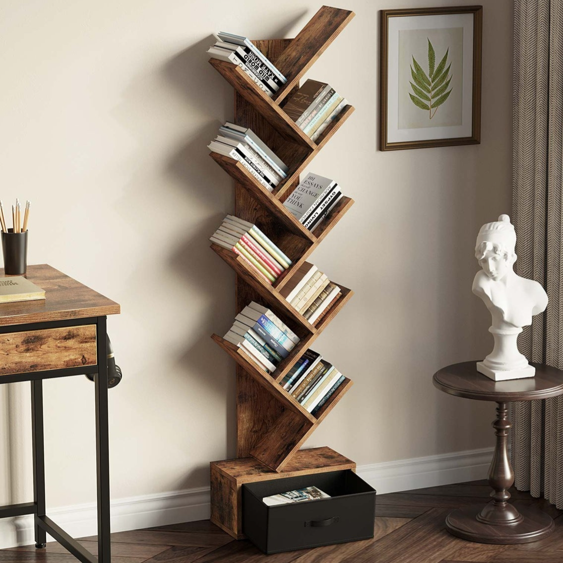 A wooden tree-style bookshelf with multiple angled shelves holds books and features a drawer at the bottom. Nearby is a small round table with a white bust sculpture and a framed leaf print on the wall.