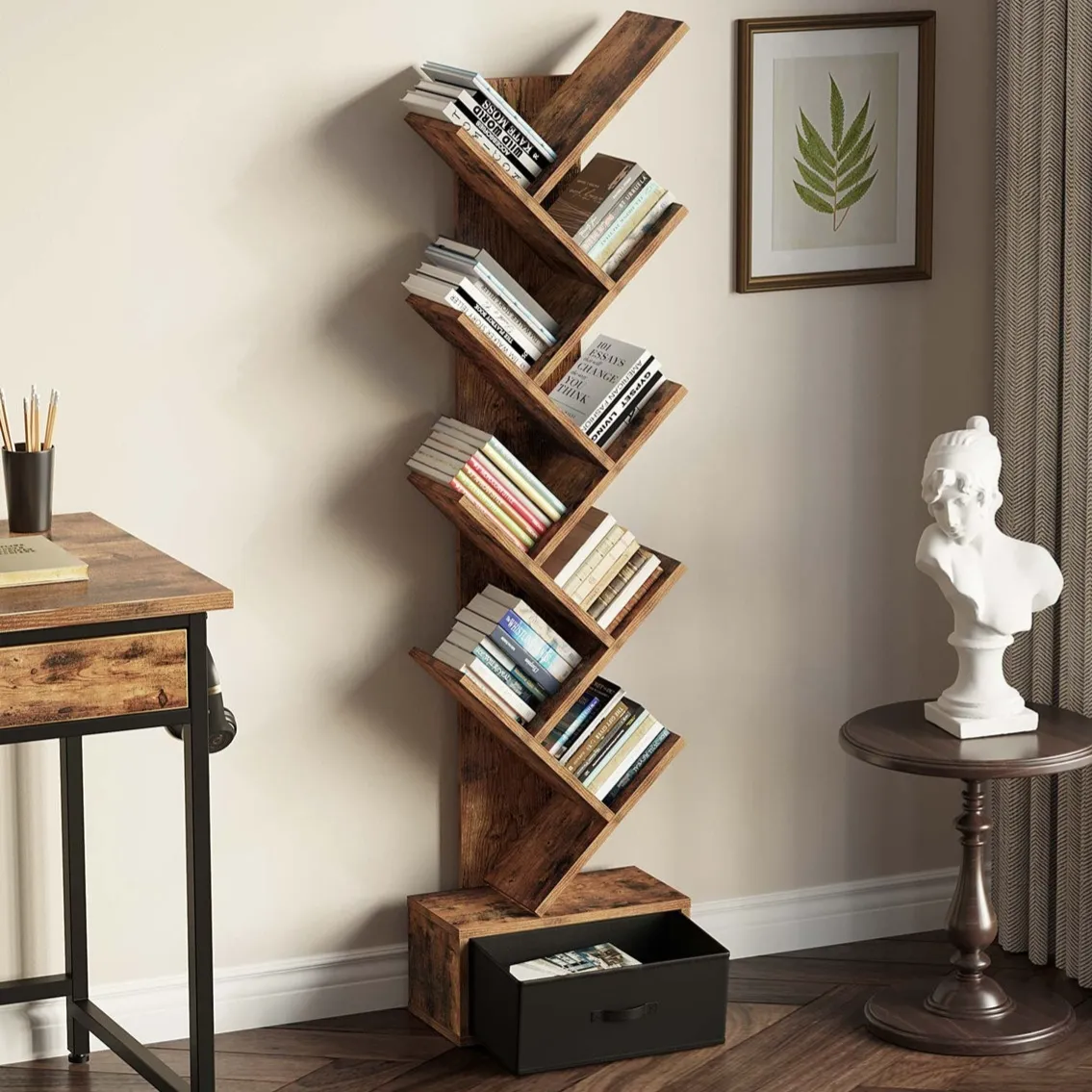 A wooden tree-style bookshelf with multiple angled shelves holds books and features a drawer at the bottom. Nearby is a small round table with a white bust sculpture and a framed leaf print on the wall.