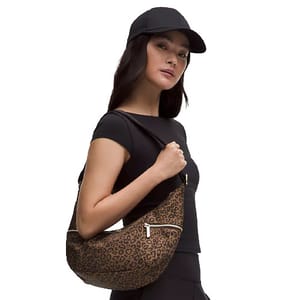 A person wearing a black cap and fitted black outfit holds a leopard print shoulder bag with zipper details.