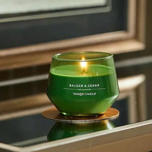 A lit green Yankee Candle with 'Balsam & Cedar' scent label is displayed on a reflective surface.