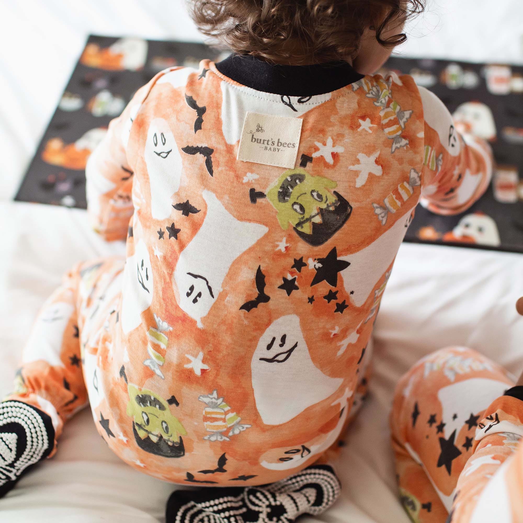 A young child is wearing Burt's Bees Baby pajamas featuring Halloween-themed prints, including ghosts, bats, and candy. The pajamas are orange with playful designs.