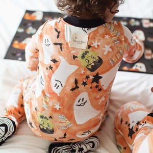 A young child is wearing Burt's Bees Baby pajamas featuring Halloween-themed prints, including ghosts, bats, and candy. The pajamas are orange with playful designs.