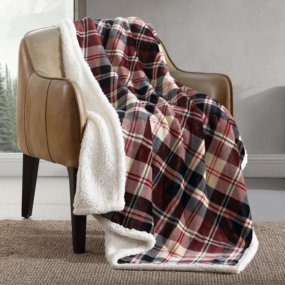 A plaid patterned sherpa throw blanket is draped over a brown leather armchair beside a small wooden side table with a plant and book, in a room with a jute rug and landscape artwork on the wall.