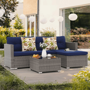 The 3pc Outdoor Sectional Set includes a gray wicker L-shaped sofa with blue cushions, two patterned pillows, and a matching glass-top coffee table. The setup is on a wooden deck with decorative string lights above.