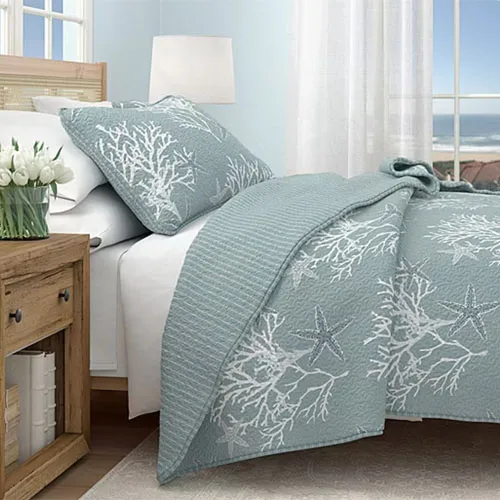 A light blue quilt and pillow with white starfish and coral designs are on a bed next to a wooden nightstand holding a bouquet of white tulips, set against a bright, beach-inspired room.
