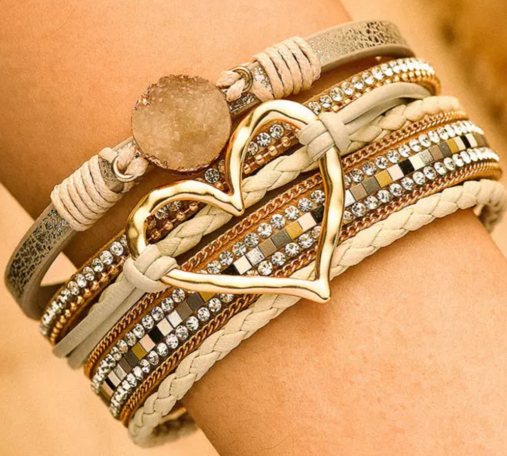 A layered bracelet features multiple bands with cream and gold tones, adorned with rhinestones, a large gold heart shape, and a circular stone accent.