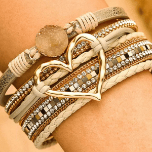 A layered bracelet features multiple bands with cream and gold tones, adorned with rhinestones, a large gold heart shape, and a circular stone accent.