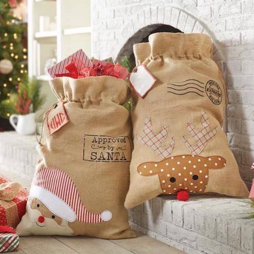 Two burlap drawstring Christmas sacks feature festive designs: one with Santa's face and the words \“Approved by Santa,\“ the other with a reindeer and plaid antlers. They are filled with wrapped gifts and positioned near a white brick hearth.