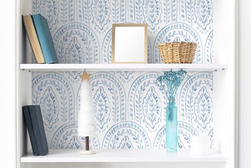 A shelf features blue books, a blank framed picture, a wicker basket, a small decorative white tree with a star, a blue vase with dried flowers, and a white candle, set against patterned wallpaper.