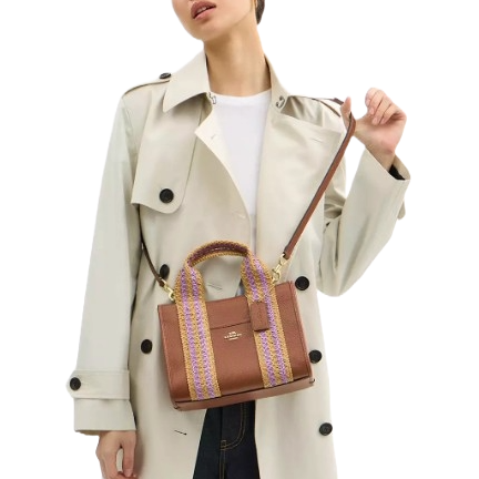 A person is wearing a light-colored trench coat and holding a brown handbag with purple and yellow woven handles and a shoulder strap.