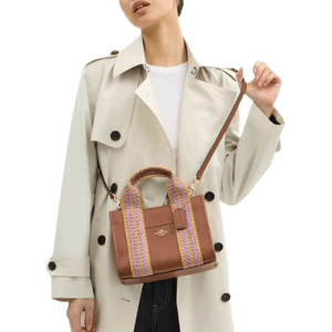 A person is wearing a light-colored trench coat and holding a brown handbag with purple and yellow woven handles and a shoulder strap.